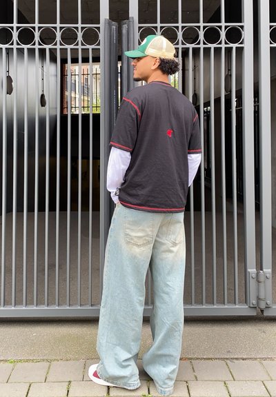 Camiseta negra de manga corta con costuras rojas, superpuesta sobre una camisa blanca de manga larga. Jeans holgados de color azul claro y una gorra verde y marrón.