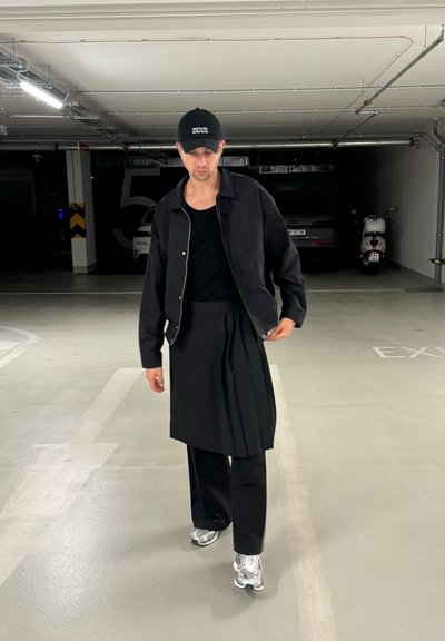 Chaqueta negra con cremallera sobre una camiseta de tirantes negra, combinada con una falda negra de tablas y pantalones anchos negros. Zapatillas plateadas completan el conjunto.