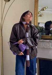 Veste aviateur en cuir marron avec manches surdimensionnées, écharpe rayée marron et blanche, sac à main violet, jean bleu à jambes larges, cadre intérieur.
