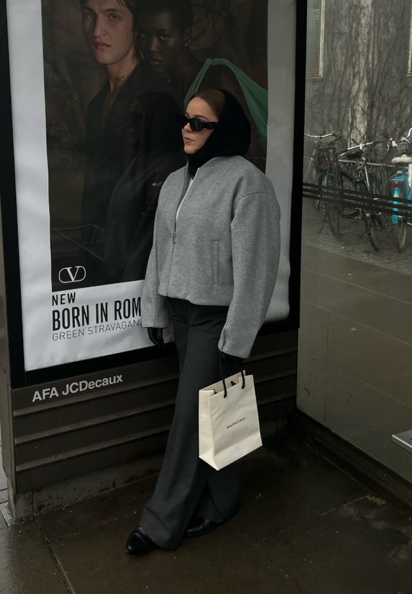 Grå kort jacka med dragkedja, svart turtleneck, vida grå byxor, håller en vit Balenciaga-väska. Urban miljö med cyklar i bakgrunden.