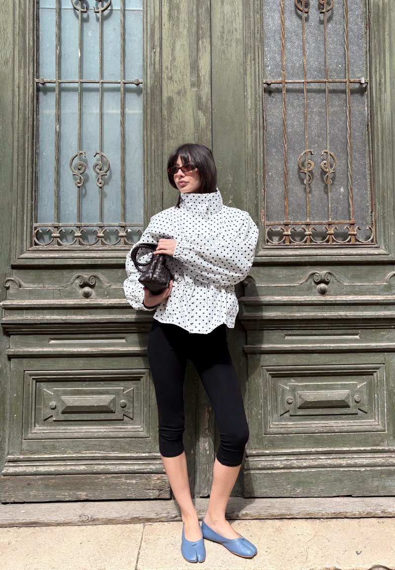 Femme aux cheveux courts et foncés, portant des lunettes de soleil, un chemisier blanc à pois, des leggings noirs courts et des chaussures bleues, tenant un petit sac noir.