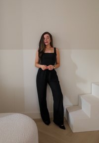 Zwarte jumpsuit met vierkante halslijn en bandjes, met een relaxte pasvorm en een koord in de taille. Geplaatst tegen een neutrale muur en met gestructureerde meubels.