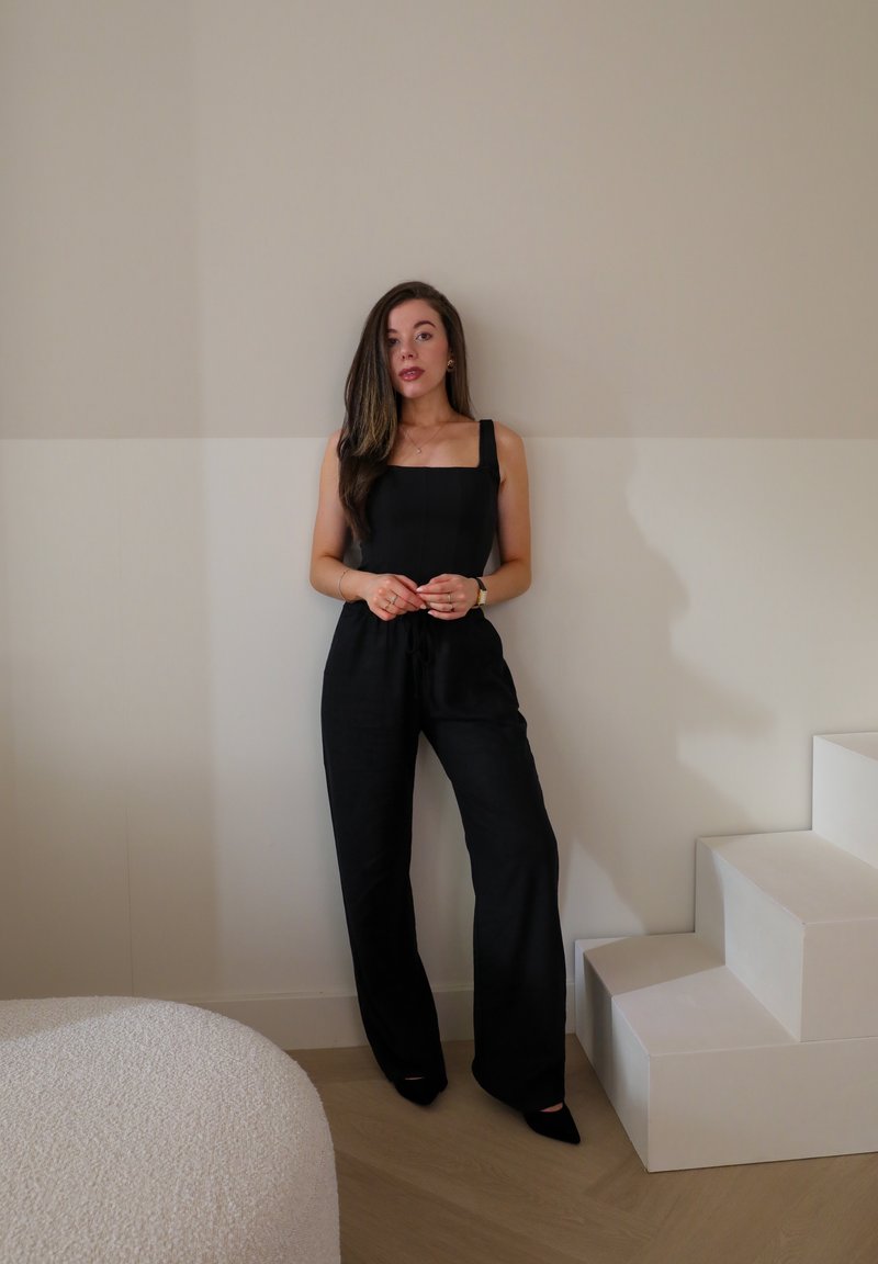Salopette noire avec décolleté carré et bretelles, présentant une coupe décontractée et une taille élastique avec cordon de serrage. Mise en scène devant un mur neutre et des meubles texturés.