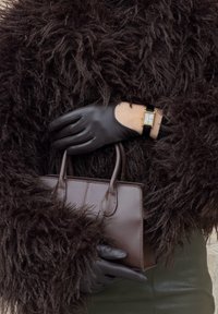 Fou de leer handtas vastgehouden door een bezette hand, met een getextureerde zwarte bontjas en een gouden polshorloge dat eronder tevoorschijn komt.
