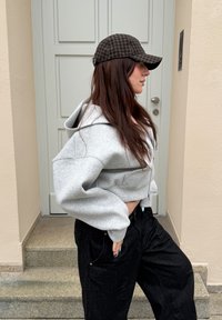 Personne portant un sweat à capuche gris, un pantalon large noir et une casquette à carreaux, se tenant de profil devant une porte blanche, main sur la hanche.