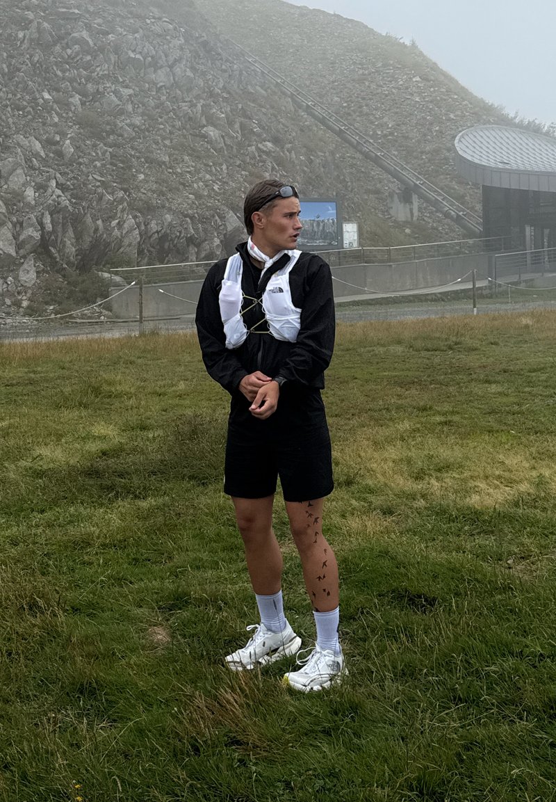 Jeune homme en veste noire et short portant un maillot de course blanc se tient sur un champ herbeux près d'une colline rocheuse et d'une structure brumeuse.