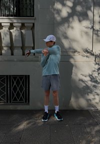 Atletische outfit met een lichtblauw shirt met lange mouwen en grijze shorts. Zwarte sneakers met blauwe accenten. Persoon die zich tegen een muur uitrekt.