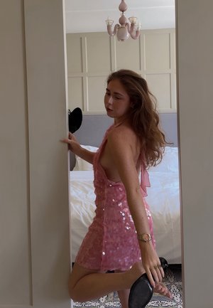 Lyserød pailletkjole med halterneck-design og åben ryg, der har en struktureret overflade. Sort højhælet sko holdes i den ene hånd.