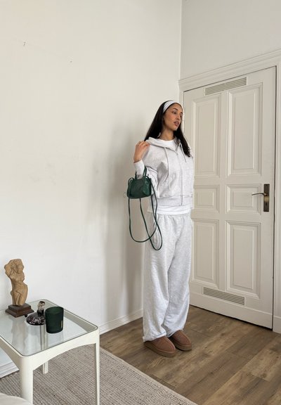 Sudadera gris recortada y pantalones deportivos a juego, combinados con pantuflas marrones y un bolso de hombro verde. Decoración minimalista en el fondo.