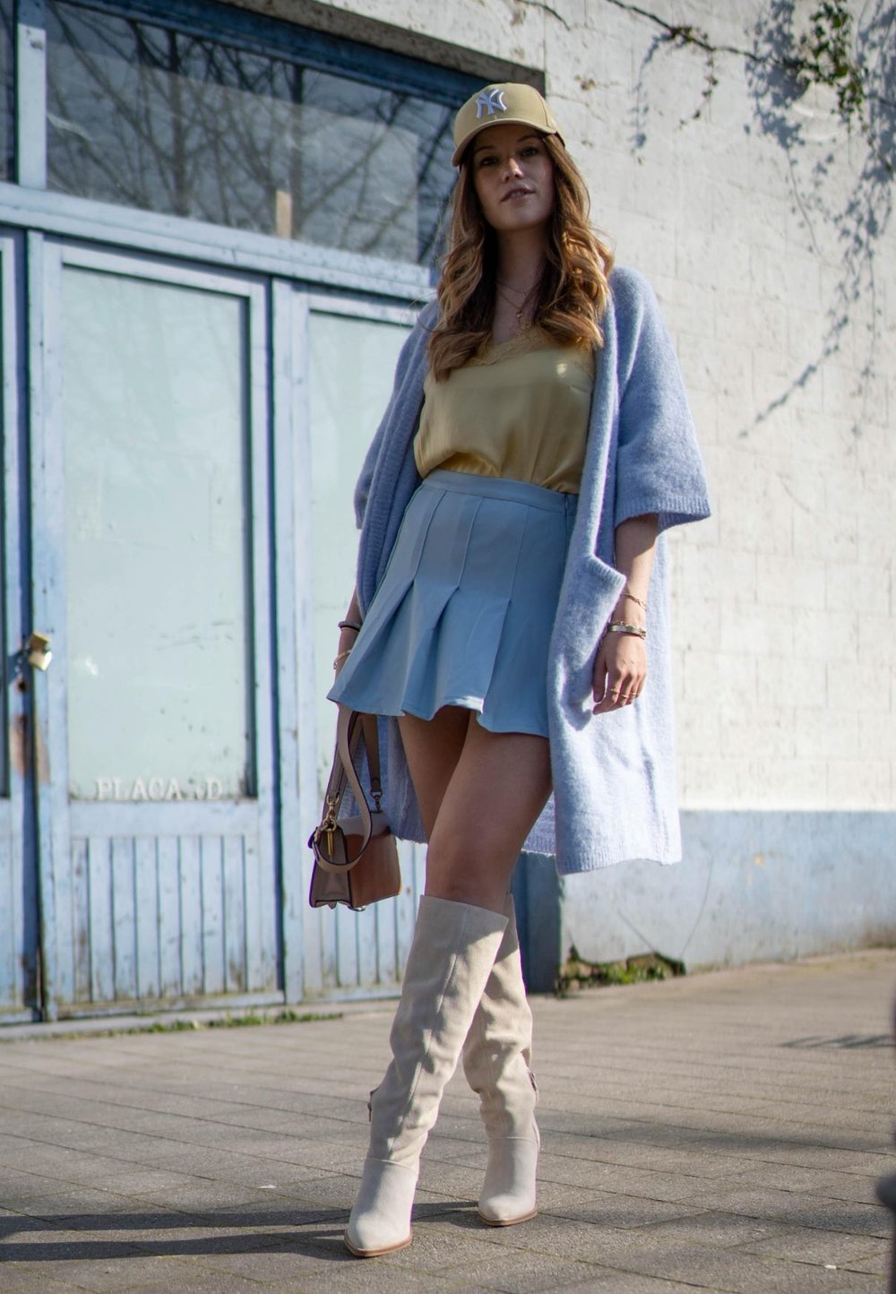 Saia mini plissada azul-claro, blusa amarela, cardigan oversized lavanda, botas beje até ao joelho e uma carteira castanha com detalhes em ouro.