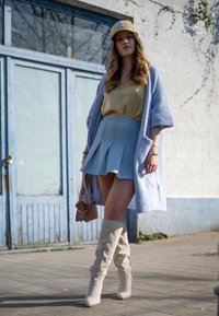 Saia mini plissada azul-claro, blusa amarela, cardigan oversized lavanda, botas beje até ao joelho e uma carteira castanha com detalhes em ouro.