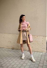 Casaco trench bege por cima de um suéter de malha rosa, combinado com uma saia bege e ténis brancos. A segurar uma bolsa de mão rosa claro. Fundo de pavimento de blocos.