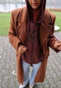 Brun uldfrakke over en vinrød hættetrøje med et leopardprintlogo, kombineret med lyseblå jeans og hvide sko på en stenspavement.