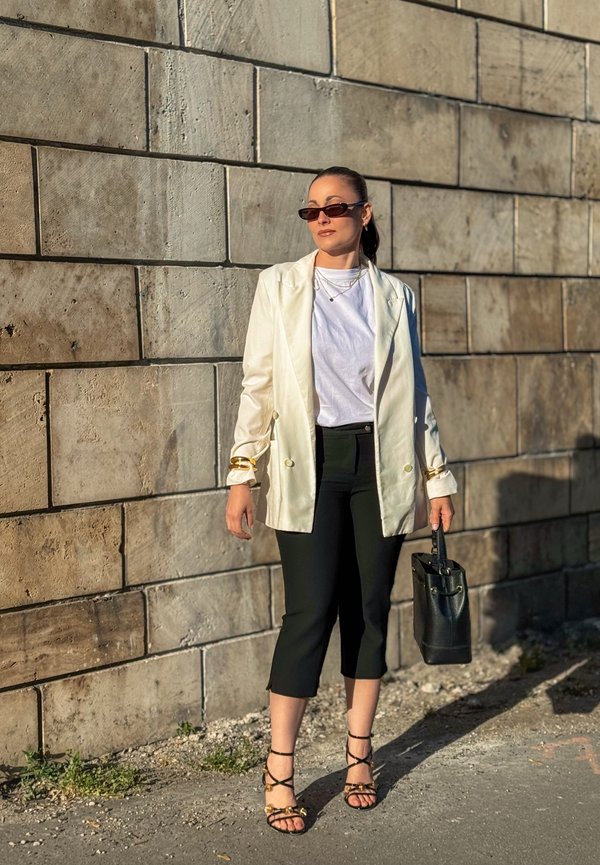 Femme portant un blazer blanc, une chemise blanche, un pantalon court noir, des lunettes de soleil et des sandales, tenant un sac à main noir, debout près d'un mur en pierre.