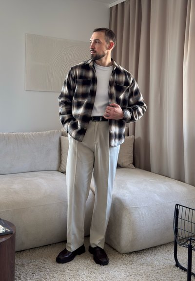 Hombre con camisa de cuadros blanca y negra sobre camiseta blanca, pantalones beige y mocasines oscuros, de pie en un salón con sofá beige y cortinas.