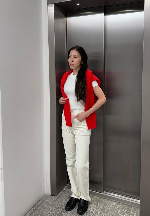 Une femme en gilet rouge et vêtue de blanc se tient devant les portes fermées d’un ascenseur.