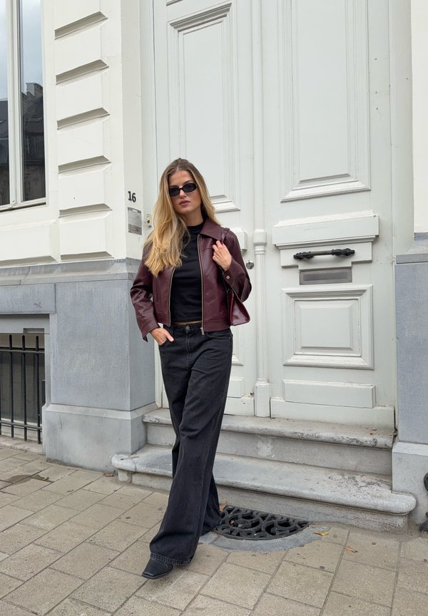 Bordo kožna jakna, crna majica i široke crne traperice. Model nosi crne sunčane naočale i stoji pored bijelih vrata sa sivoj obrubom.