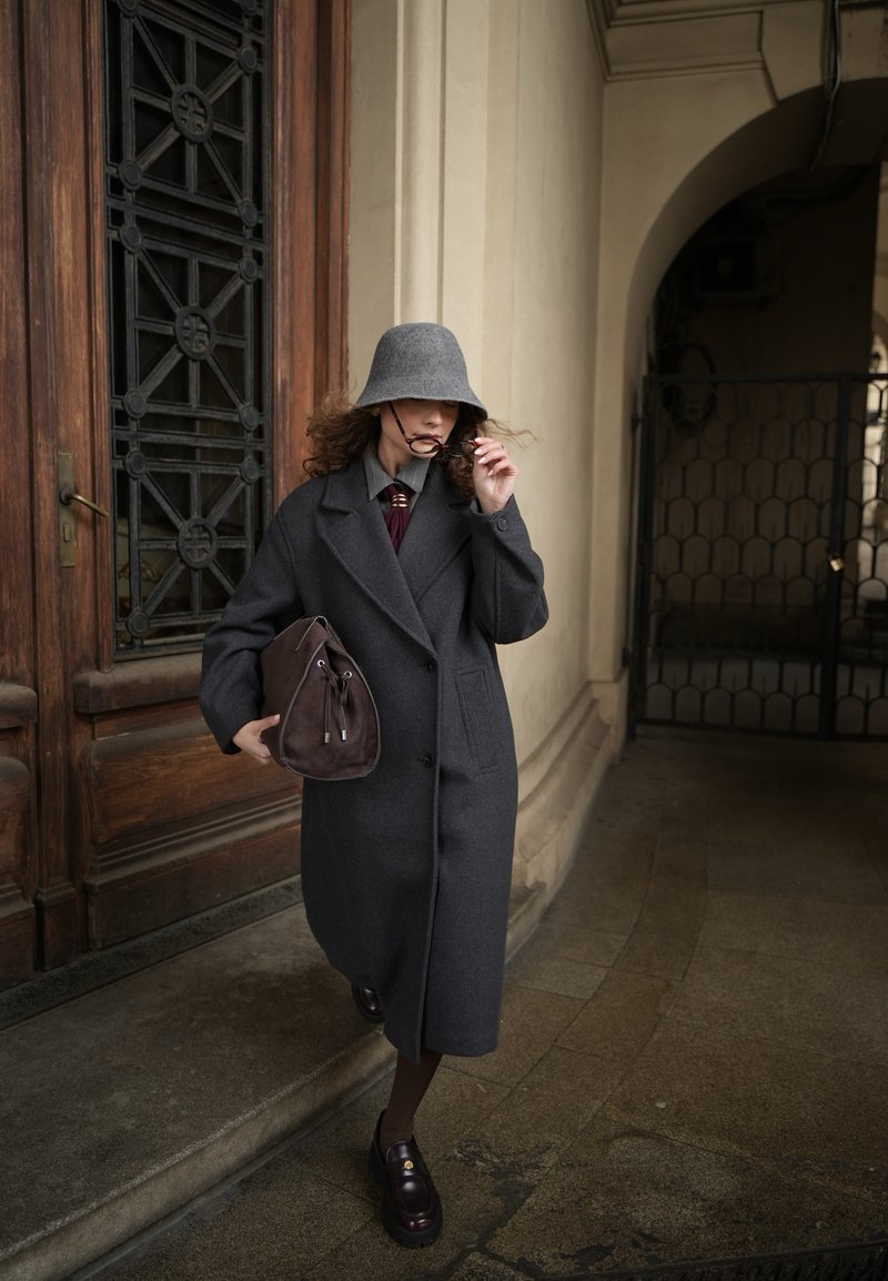 Femme en manteau gris foncé et chapeau seau tenant un sac marron et ajustant ses lunettes de soleil, debout près d'une porte en bois dans un couloir voûté.