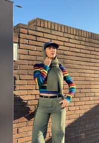 Γυναίκα με πουλόβερ ριγέ με ουράνιο τόξο και παντελόνι ελιάς, στέκεται μπροστά σε τοίχο από τούβλα, φορώντας μεγάλο κασκόλ ελιάς και μπλε σκουφί.