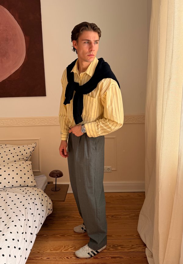 Jonge man met een geel gestreept overhemd, grijze broek, zwarte trui over de schouders, witte sneakers, staand bij het bed en het gordijn in de slaapkamer.