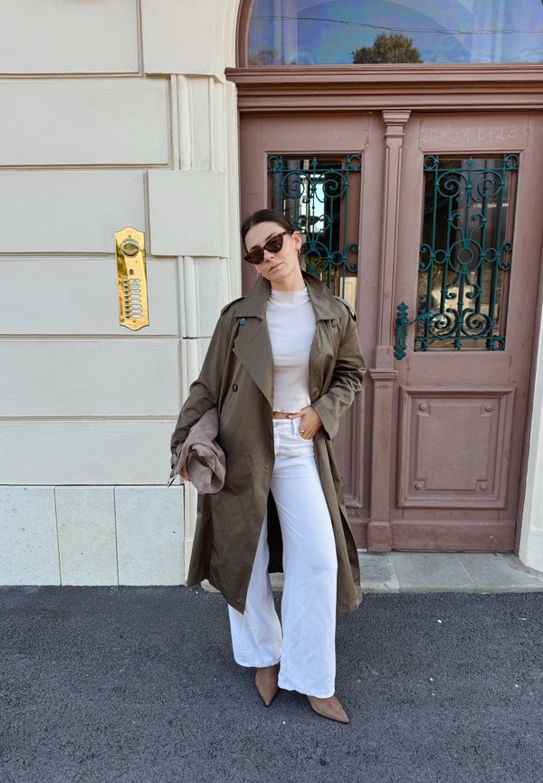 Mujer con gafas de sol, abrigo marrón, top blanco y pantalones anchos blancos, junto a una pared beige y una puerta marrón con rejillas metálicas.