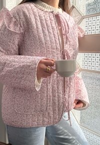 Lyserød quiltet jakke med blomstermønster, flæseærmer og båndlukning. Båret over en cremefarvet sweater, holdt med en hvid kop.