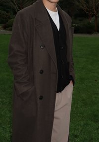 Manteau long en laine mélangée marron avec de gros boutons, superposé à un cardigan noir, porté avec un pantalon beige clair. Environnement extérieur avec de l'herbe.