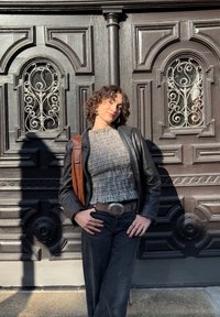 Zwarte leren jas over een zwart-wit geblokte top, gecombineerd met donkere jeans. Een bruine tasband is zichtbaar. Ornate houten achtergrond.