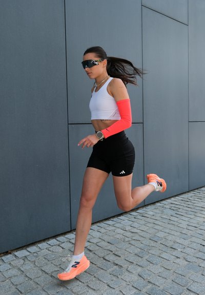Corredora femenina vestida con una camiseta sin mangas blanca, pantalones cortos negros y manguitos naranjas. Lleva unas gafas de sol negras y zapatillas de running naranjas.
