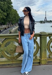 Giacca di pelle nera, top corto beige, jeans ampi blu, borsa marrone, occhiali da sole oversize; in piedi accanto a un fiume con la Torre Eiffel sullo sfondo.