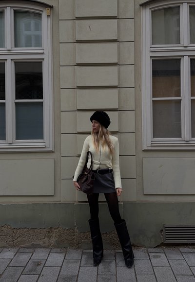 Joven mujer con sombrero negro, suéter blanco, falda gris y botas negras de pie en una acera frente a un edificio beige con dos ventanas.