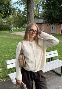 Crème-kleurige, gestructureerde cardigan met knopen, gecombineerd met donkere broeken. Een kleine bruine tas hangt over de schouder en bevat een tijdschrift.