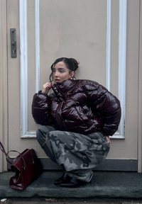 Burgundy glossy puffer jacket with high collar, paired with grey camouflage trousers and black ankle boots. A burgundy handbag rests on the ground.