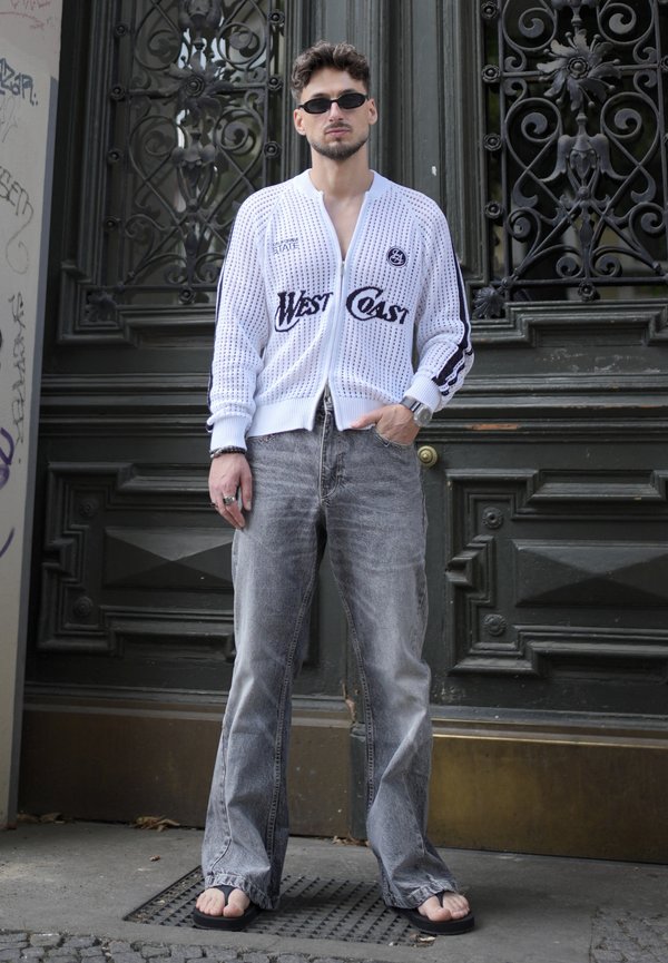 Mann mit Sonnenbrille, weißem Reißverschluss-Pullover mit dem Text "West Coast", grauen Jeans und schwarzen Flip-Flops steht vor einer verzierten dunklen Tür.