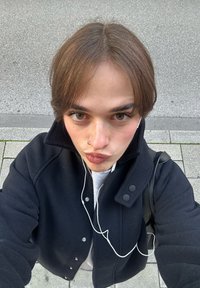Čierna bunda s okrúhlym golierom, sivý sveter pod ňou, viditeľné slúchadlá, svetlohnedé vlasy, stojaci na chodníku s roztrúsenou trávou.
