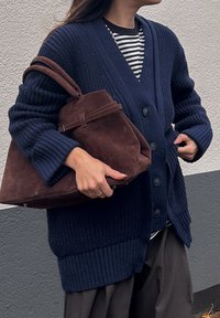 Cardigan em ponto azul marinho com tecido texturizado, fecho de botão, mangas largas; modelo segurando uma mala de camurça castanha com uma forma estruturada.
