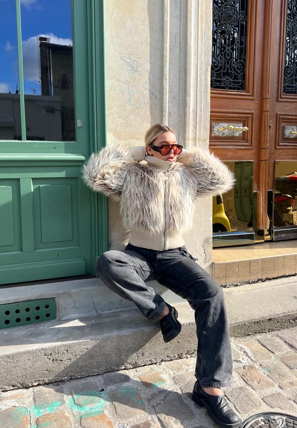 Chaqueta de piel sintética con una base crema y mangas texturizadas y esponjosas, combinada con unos pantalones oscuros de corte holgado y zapatos negros. La luz brillante del sol ilumina la escena.