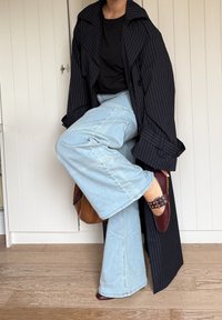 Dlhý námořnicky modrý kabát s tenkými prúžkami, čierne tričko, svetlomodré rifle s širokými nohavicami, bordové topánky s cvočkovým remienkom, hnedá kabelka, pozadie z drevenej podlahy.