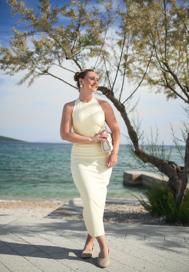 Jeune femme en robe jaune clair et chaussures beiges souriant près de la mer à côté d’un arbre par une journée ensoleillée.