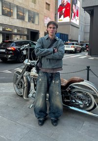Jaqueta de denim e camisa de xadrez com jeans largos e desgastados. Ao lado de uma motocicleta prateada com banco de couro marrom e design intrincado nas rodas.