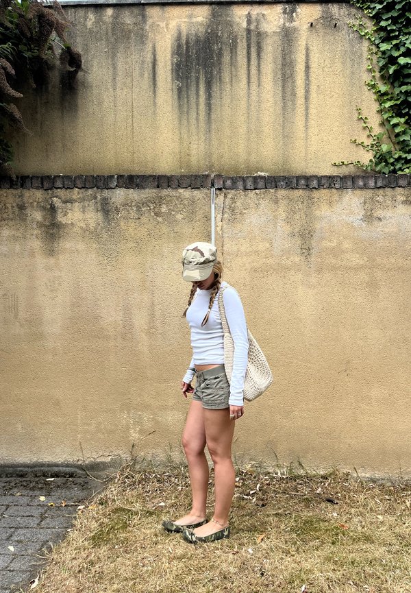 Joven mujer con el cabello trenzado, vestida con una camiseta blanca de manga larga, pantalones cortos de camuflaje, gorra y zapatos, de pie contra una pared beige desgastada.