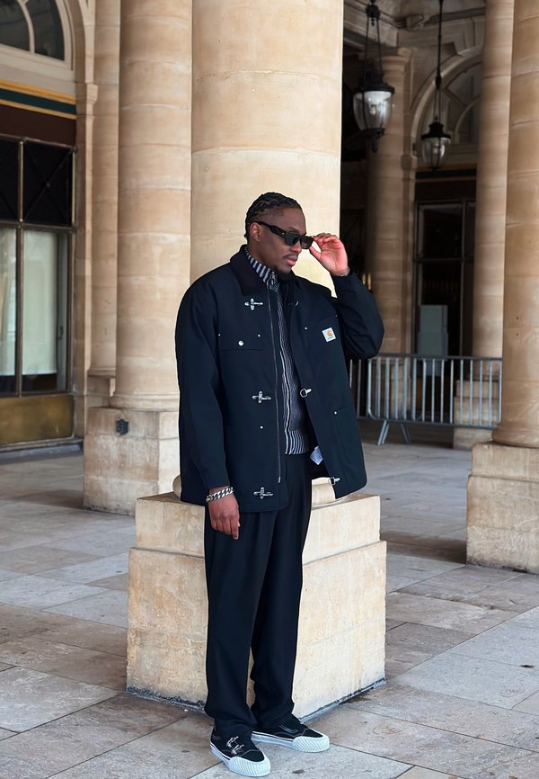 Hombre con chaqueta negra, camisa de rayas y zapatillas, ajustándose las gafas de sol, de pie junto a una columna de piedra en un área exterior cubierta.