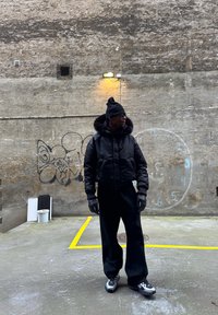 Fekete, rövidített pehelykabát szőrm�ével díszítve, fekete sapka, fekete kesztyű, és bő, fekete nadrág. Szürke és fekete részletekkel ellátott tornacipők. Városi háttér.