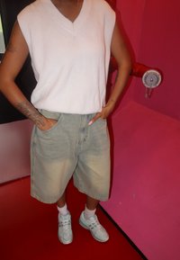 Top bianco senza maniche, con scollatura alta, abbinato a pantaloni corti in denim chiaro e sneakers azzurre. Lo sfondo presenta pareti rosa e nere.