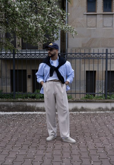Hombre con camisa azul, pantalones beige, suéter negro colgado sobre los hombros, gorra y gafas de sol, de pie en la calle cerca de un árbol en flor y una valla de metal.