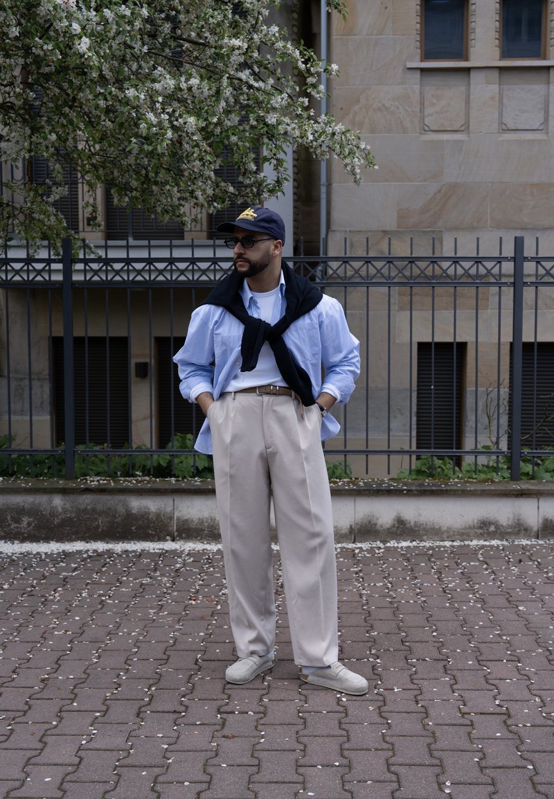 Homme portant une chemise bleue, un pantalon beige, un pull noir posé sur les épaules, une casquette et des lunettes de soleil, debout dans la rue près d’un arbre en fleurs et d’une clôture en métal.