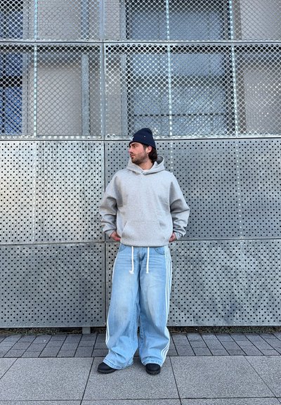 Sudadera con capucha gris, jeans de pierna ancha azul claro con rayas blancas, calzado negro, de pie contra una pared de metal perforada.