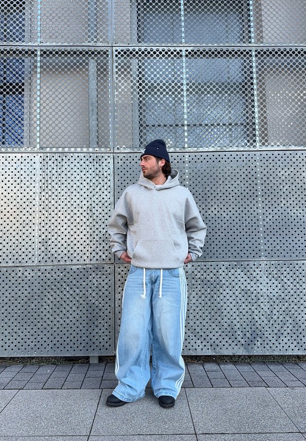 Felpa con cappuccio grigia, jeans a gamba larga blu chiaro con strisce bianche, calzature nere, in piedi contro un muro di metallo perforato.
