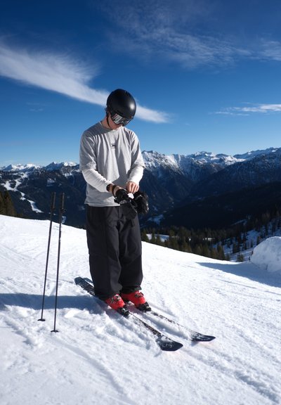 Esquiador con casco negro y gafas ajusta sus guantes en una pendiente nevada, con bastones de esquí plantados a su lado, montañas nevadas y cielo azul en el fondo.