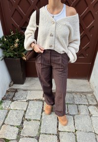 Cardigan beige en tricot avec trois boutons, associé à un pantalon rayé brun. Chaussures à enfiler couleur fauve complètent la tenue. En arrière-plan, une plante.